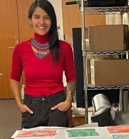 Smiling human with long dark hair wearing a red top and colorful necklace, standing behind tables with papers and boxes in a room with metal shelving.