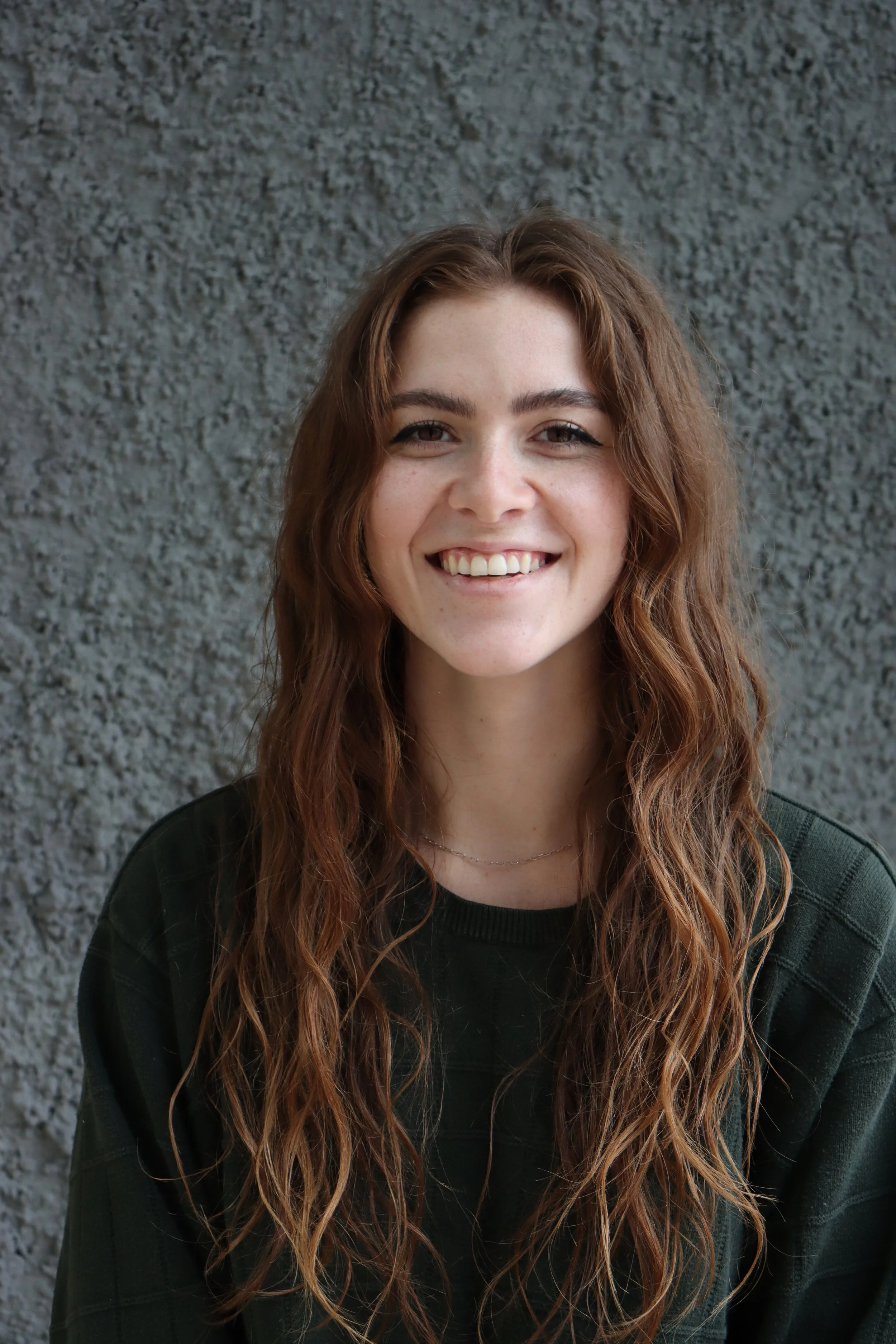 A young person with long, wavy reddish-brown hair smiling at the camera, wearing a dark green sweatshirt, standing against a textured gray wall.
