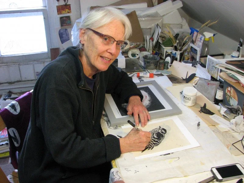 An person with glasses and white hair, sitting at a cluttered art desk, drawing a black and white illustration of an owl on paper with a pen.