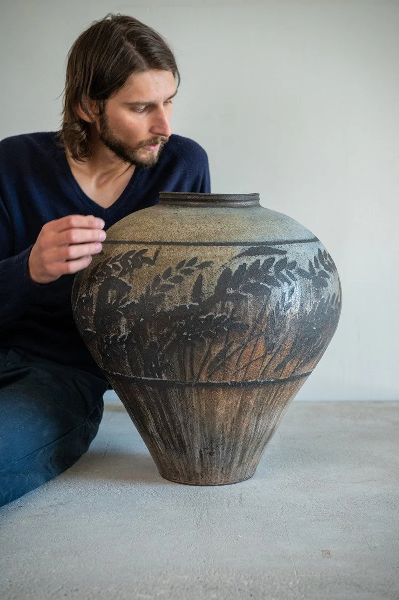 A person with long hair and beard inspecting a large ceramic jar with decorative dark leaf patterns, sitting on a plain light-colored floor against a plain wall.