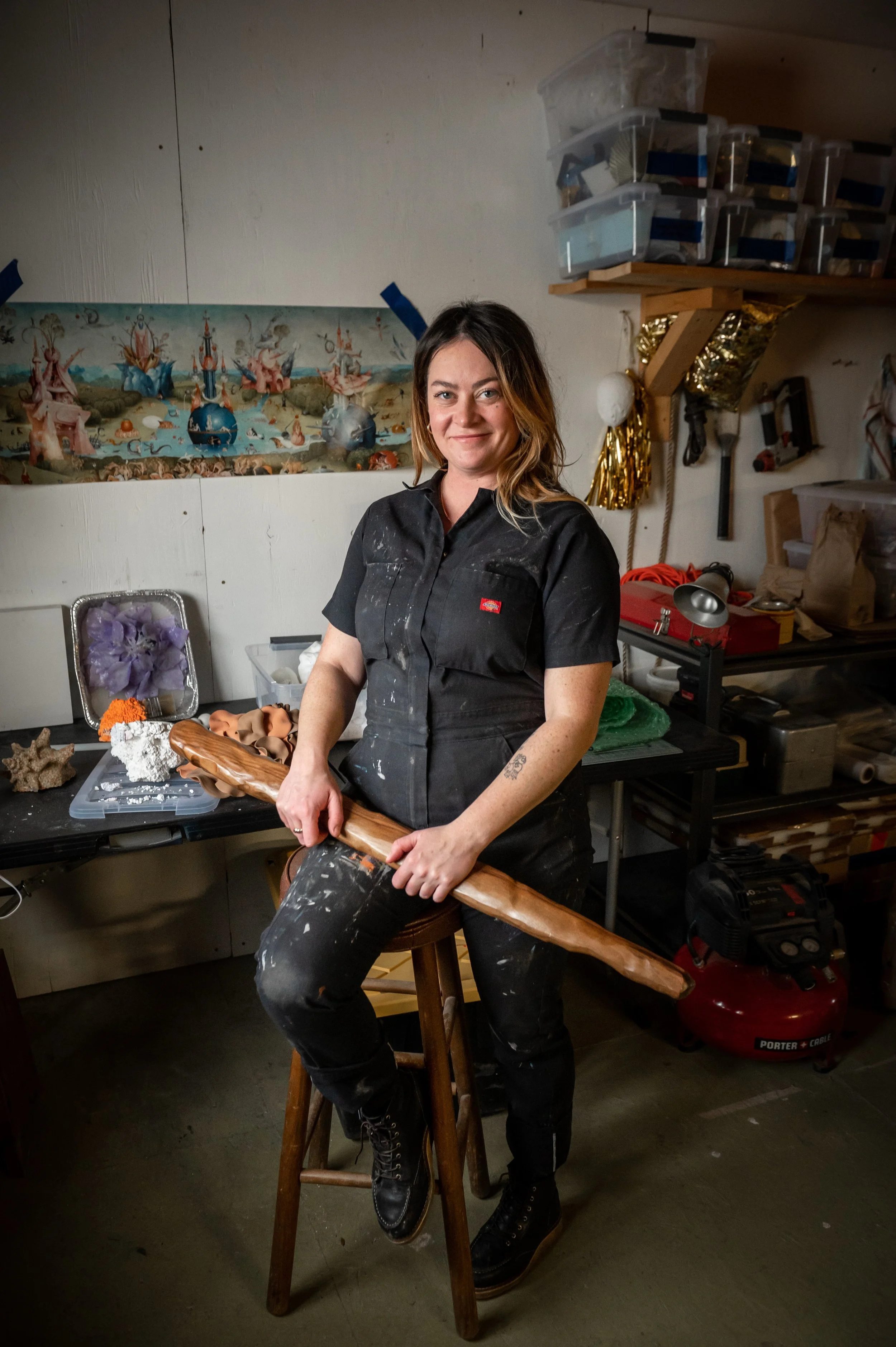 A person in black work clothes holding a wooden sword, standing in a cluttered workshop with art and craft supplies.