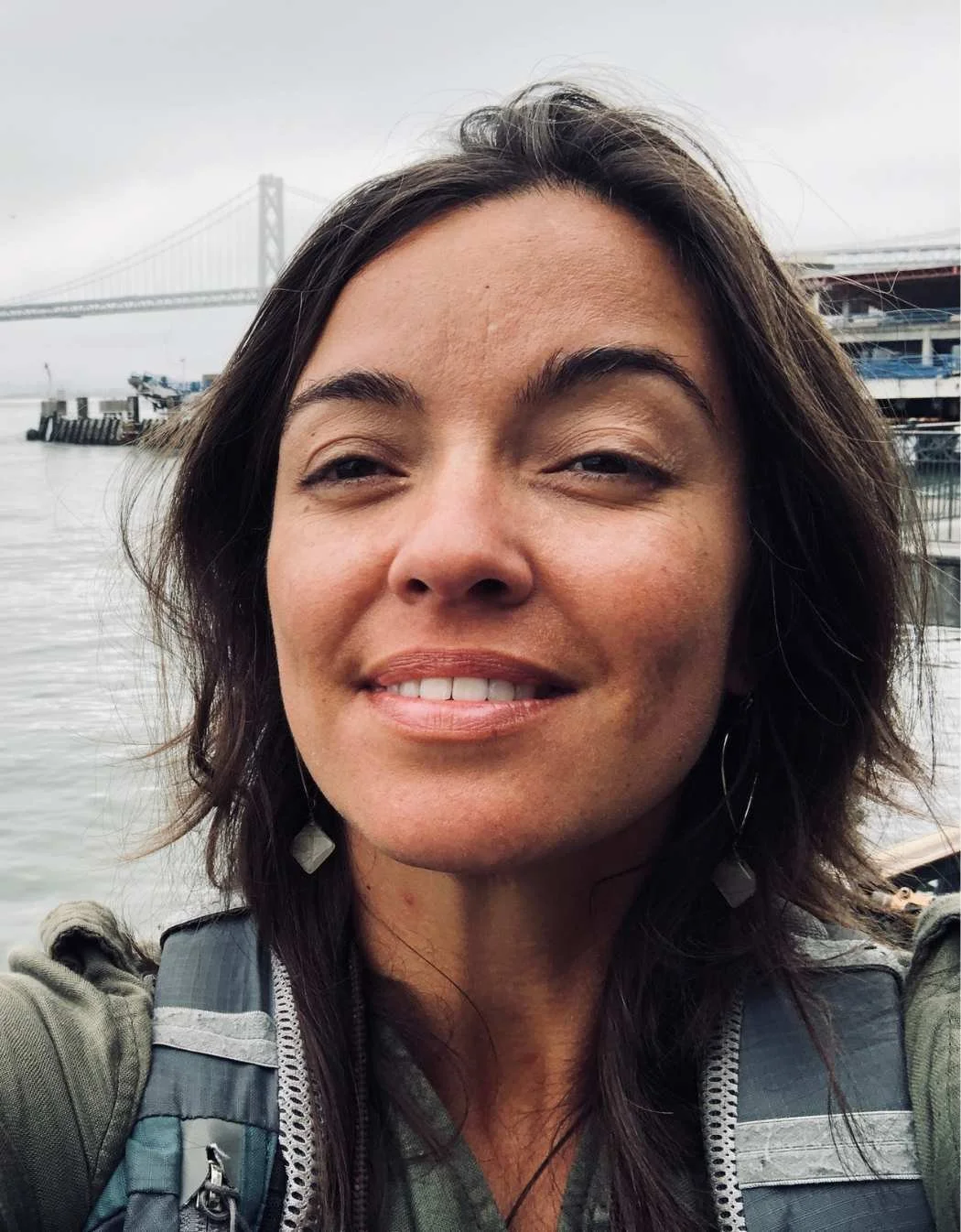 Close-up of a person smiling outdoors with a bridge and water in the background.
