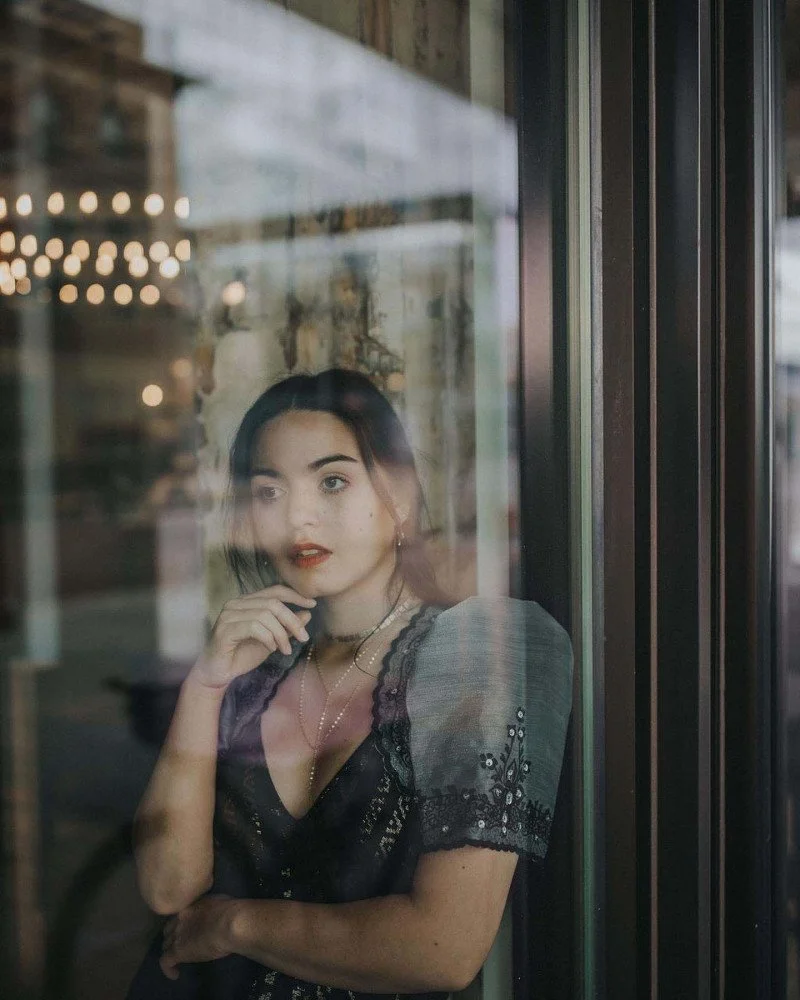 A person with dark hair and red lipstick looking out a window with reflections and bokeh lights in the background.