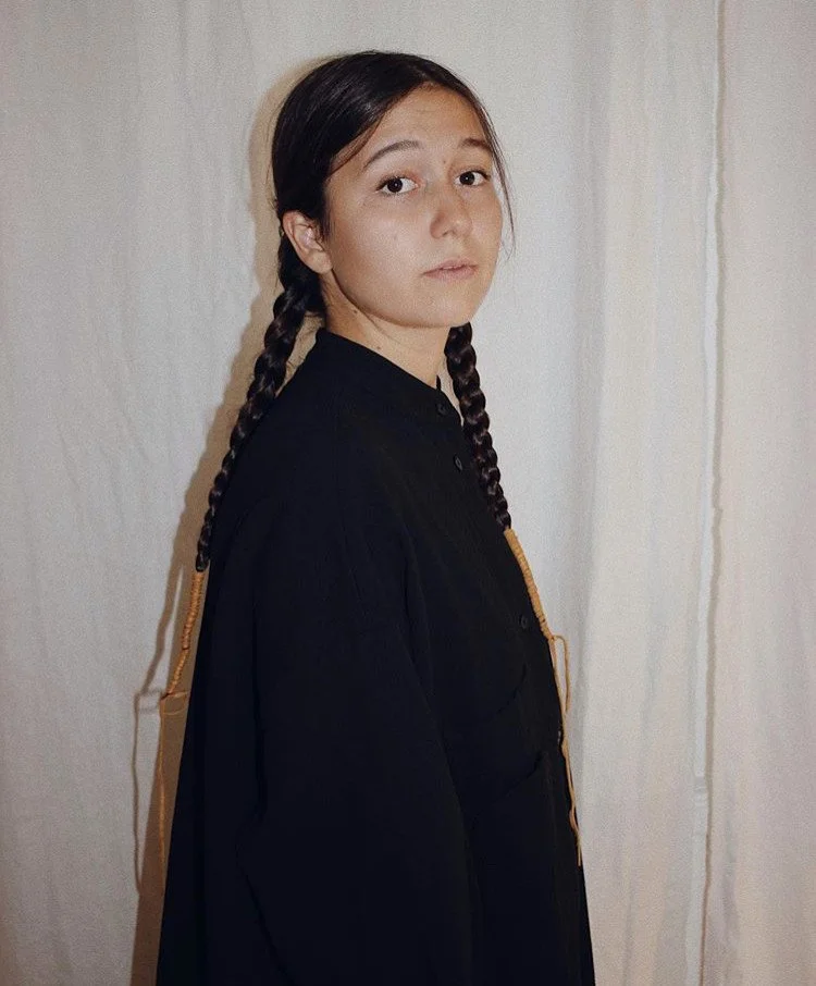 A young person with long dark hair in two braids stands sideways against a plain off-white wall, looking over her shoulder at the camera, wearing a black shirt or jacket.