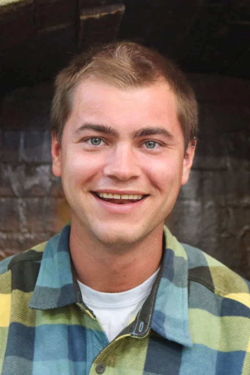 A young person smiling, wearing a colorful plaid shirt, with a brick wall background.