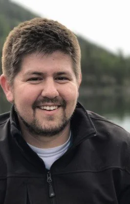 A person  with short dark hair and a beard smiling outdoors near a lake with trees in the background.