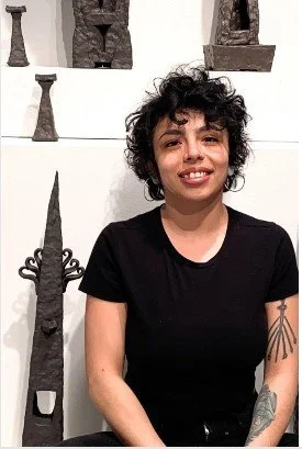 Young person with curly hair smiling, sitting in front of a wall decorated with abstract sculptures.