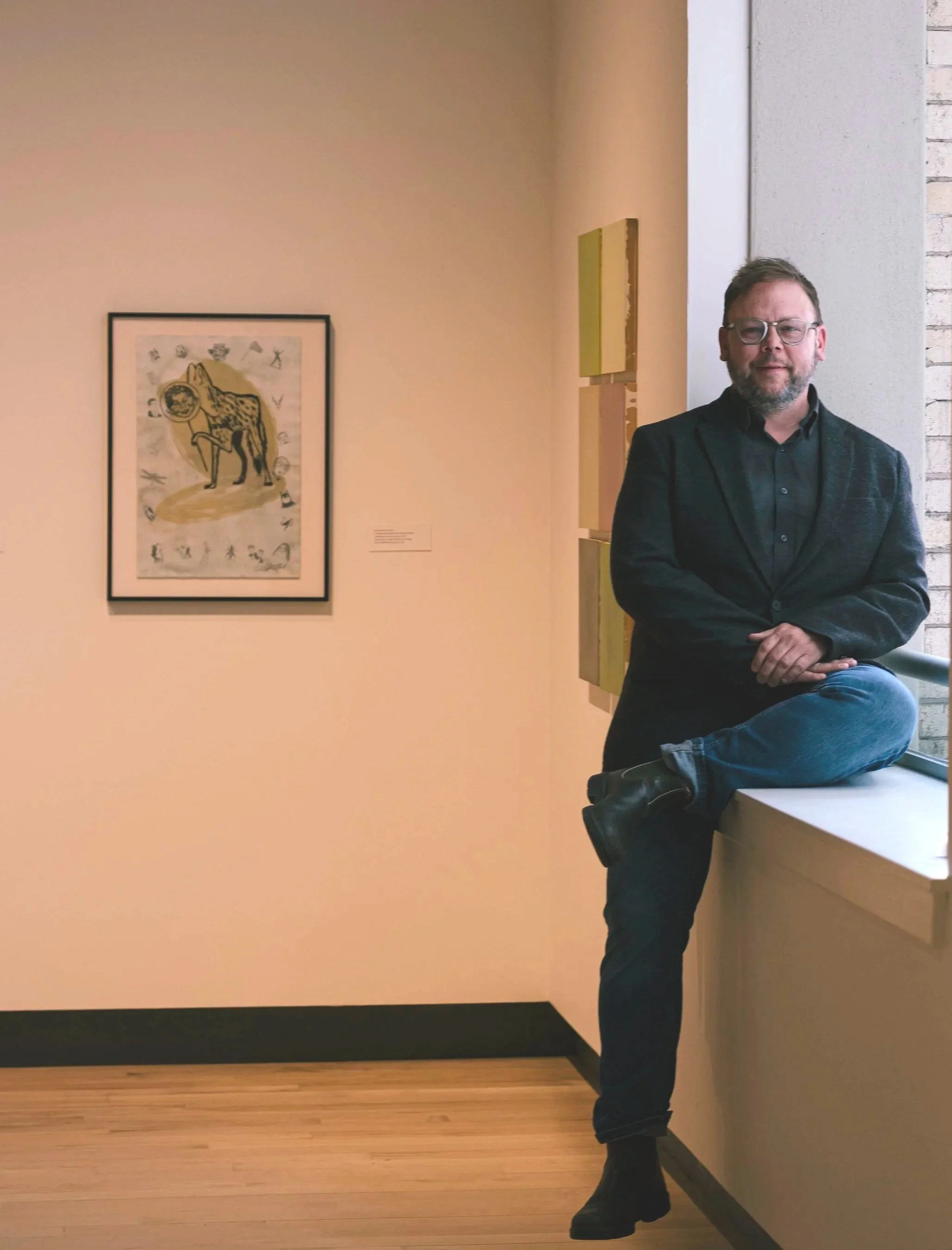 A person with glasses and a beard wearing a red plaid shirt, smiling, with art or photographs on the wall in the background.