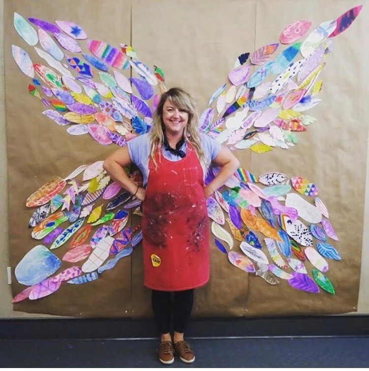 Person standing with hands on hips, wearing a red apron, in front of a large wall art of butterfly wings made of colorful paper or fabric cutouts.