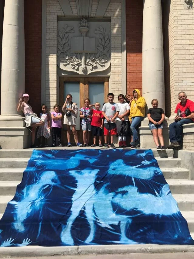 A pregnant woman in a milk bath surrounded by pink, white, and cream-colored flowers and petals, holding a white flower close to her belly.