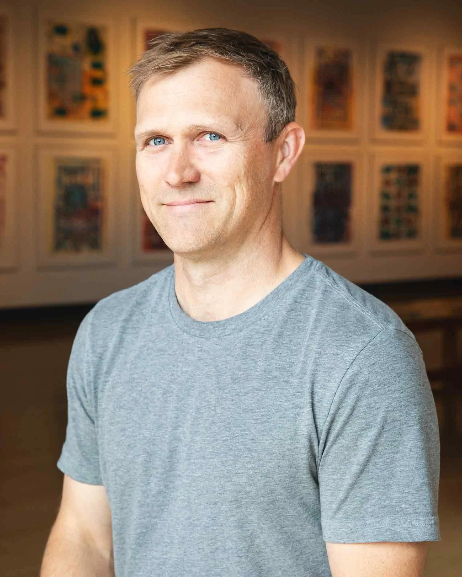 A person with short light brown hair and blue eyes, wearing a gray t-shirt, smiling slightly, standing in a room with artwork on the wall behind him.