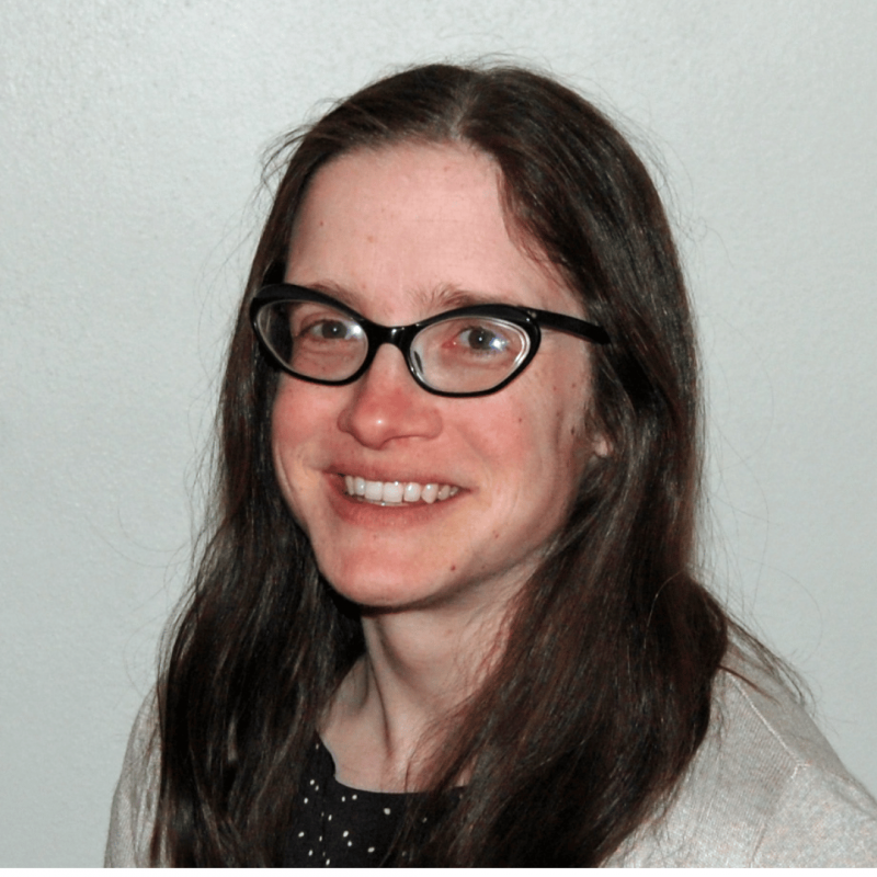 A person with long brown hair, wearing glasses, smiling, against a plain white wall.