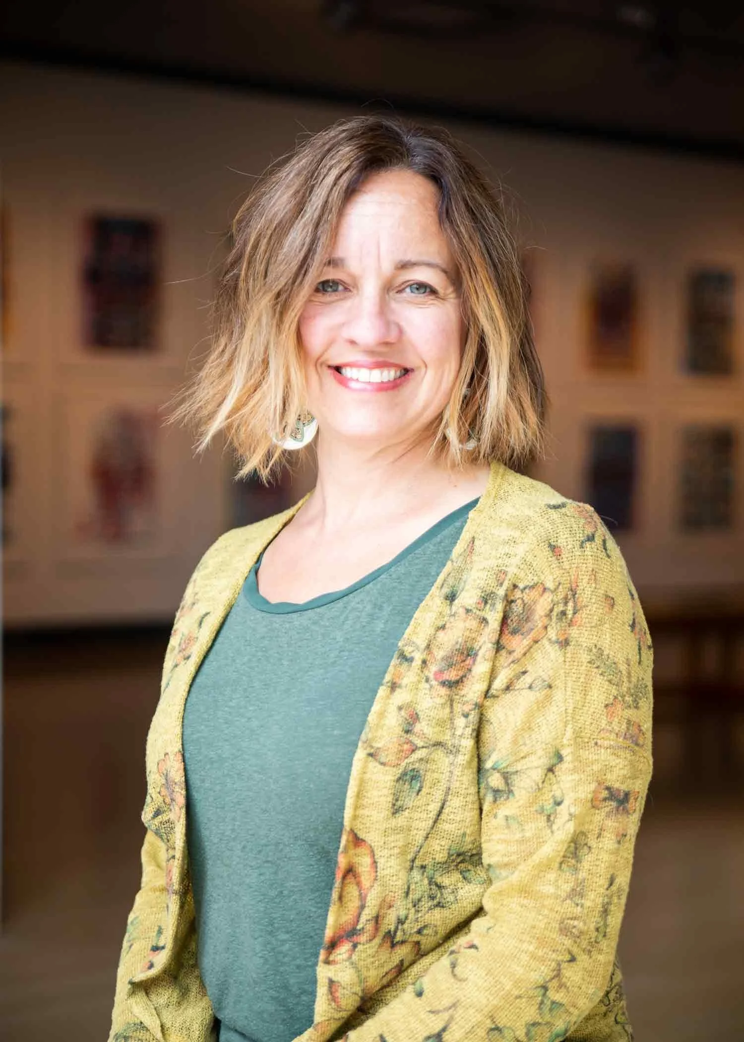 A person with shoulder-length blond hair, smiling, wearing a green shirt and a yellow floral blazer, standing indoors with a blurred background of framed artwork.
