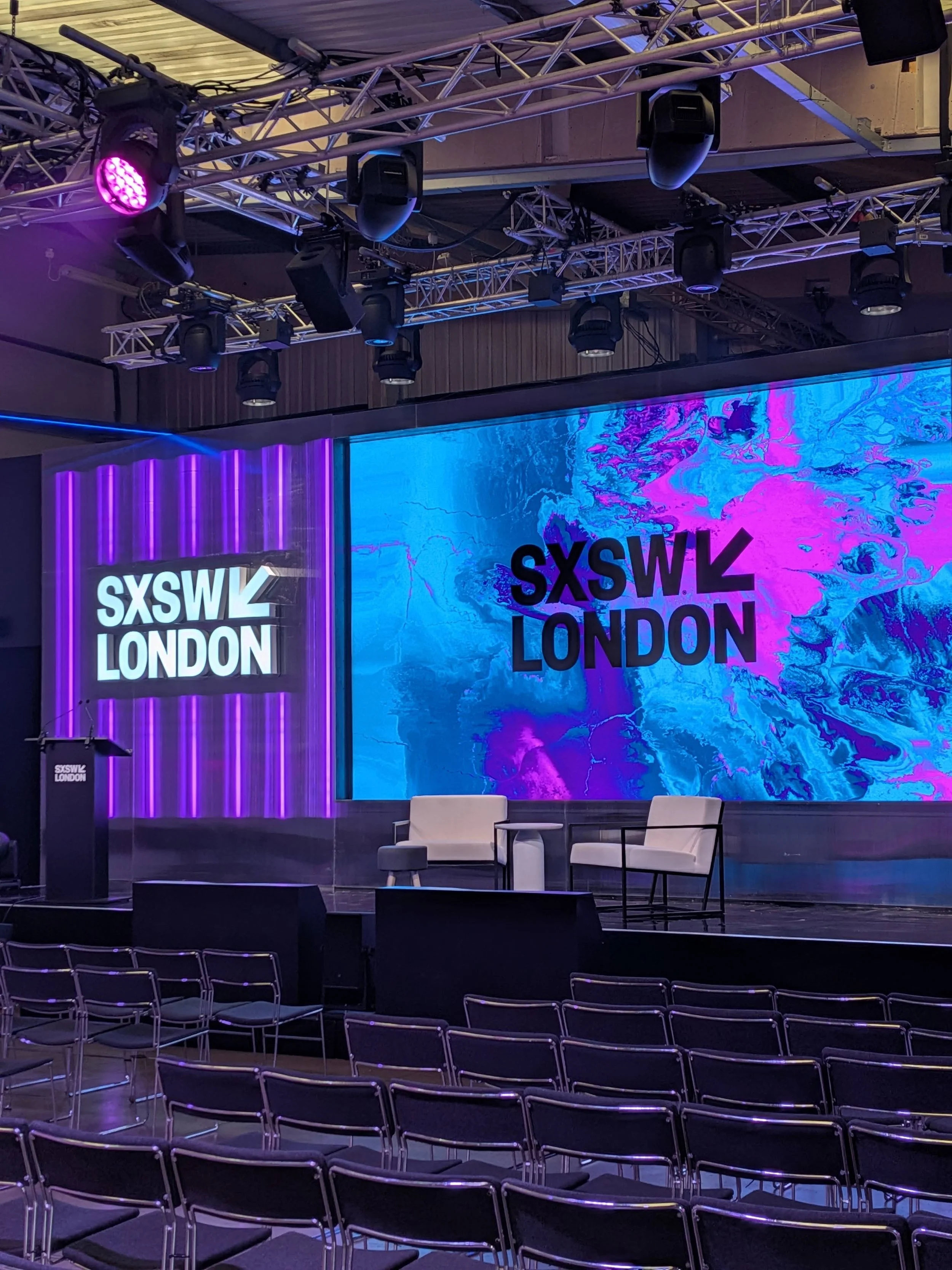 Empty stage with chairs, large screen displaying 'SXSW London,' and theater lighting equipment overhead at SXSW London event