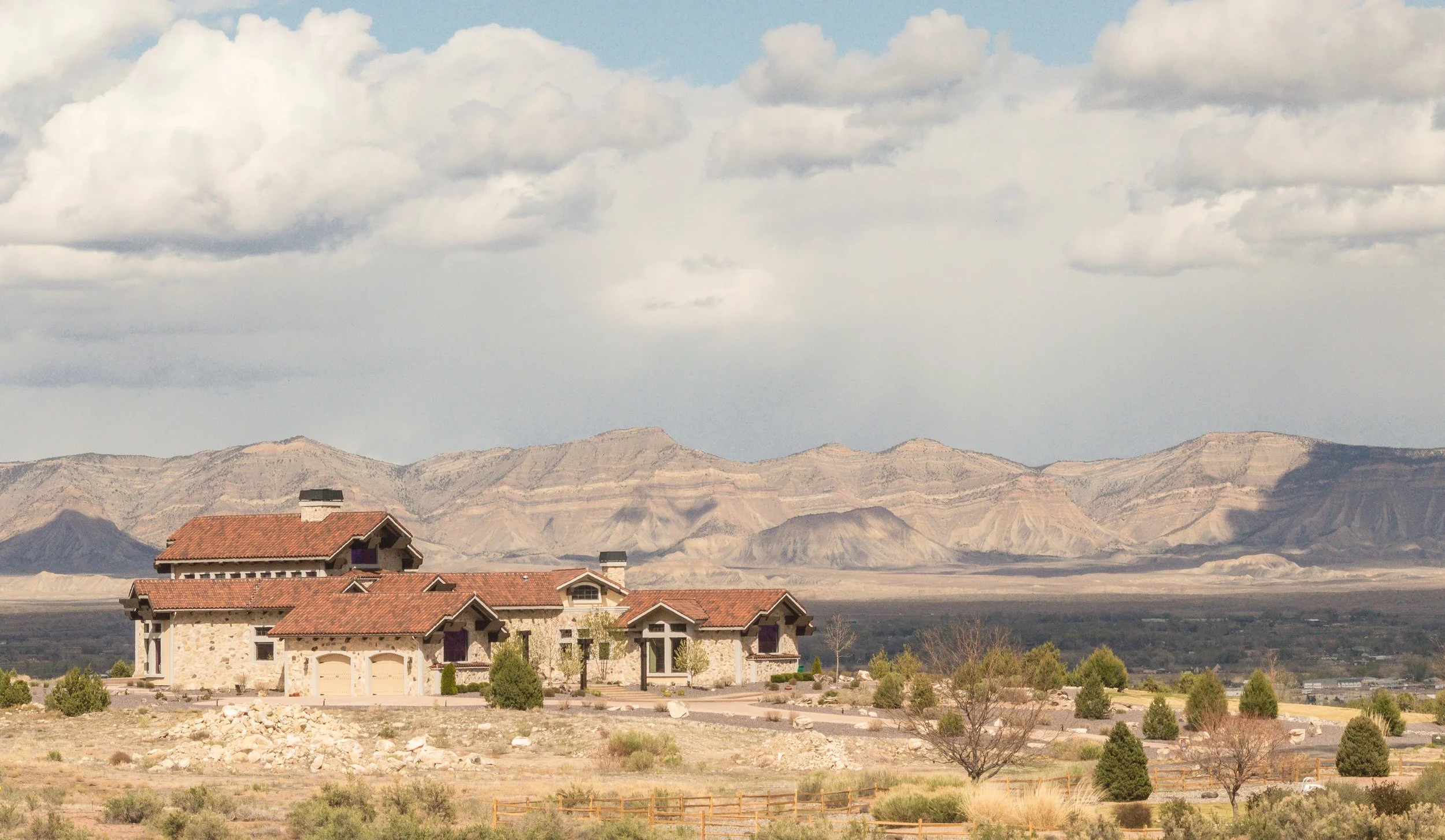 A residential area with houses featuring red tile roofs in a desert landscape, with mountains and cloudy sky in the background.