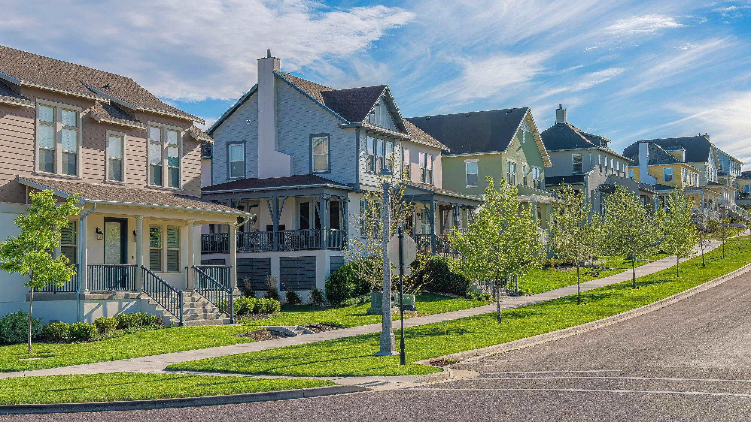 A row of colorful modern houses with porches, surrounded by green lawns, trees, and a sidewalk, under a partly cloudy sky.