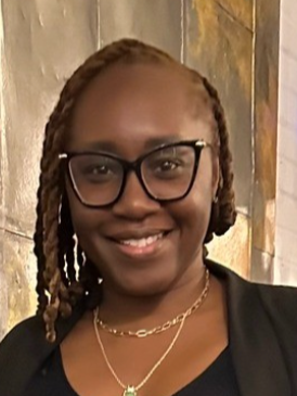 Close-up of a smiling woman with glasses and braided hair, wearing a black top and layered necklaces.