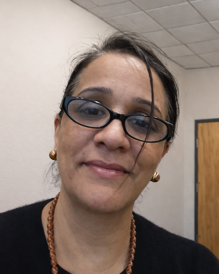 Close-up of a woman wearing glasses, earrings, and a necklace, smiling in an indoor setting.