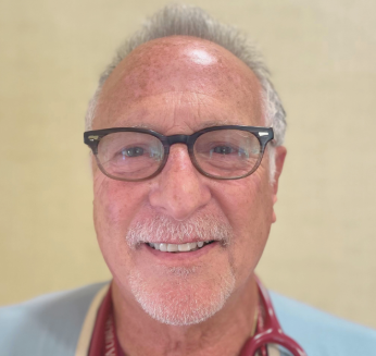 A smiling older man with glasses, white hair, and a beard, wearing a medical stethoscope around his neck against a light-colored background.
