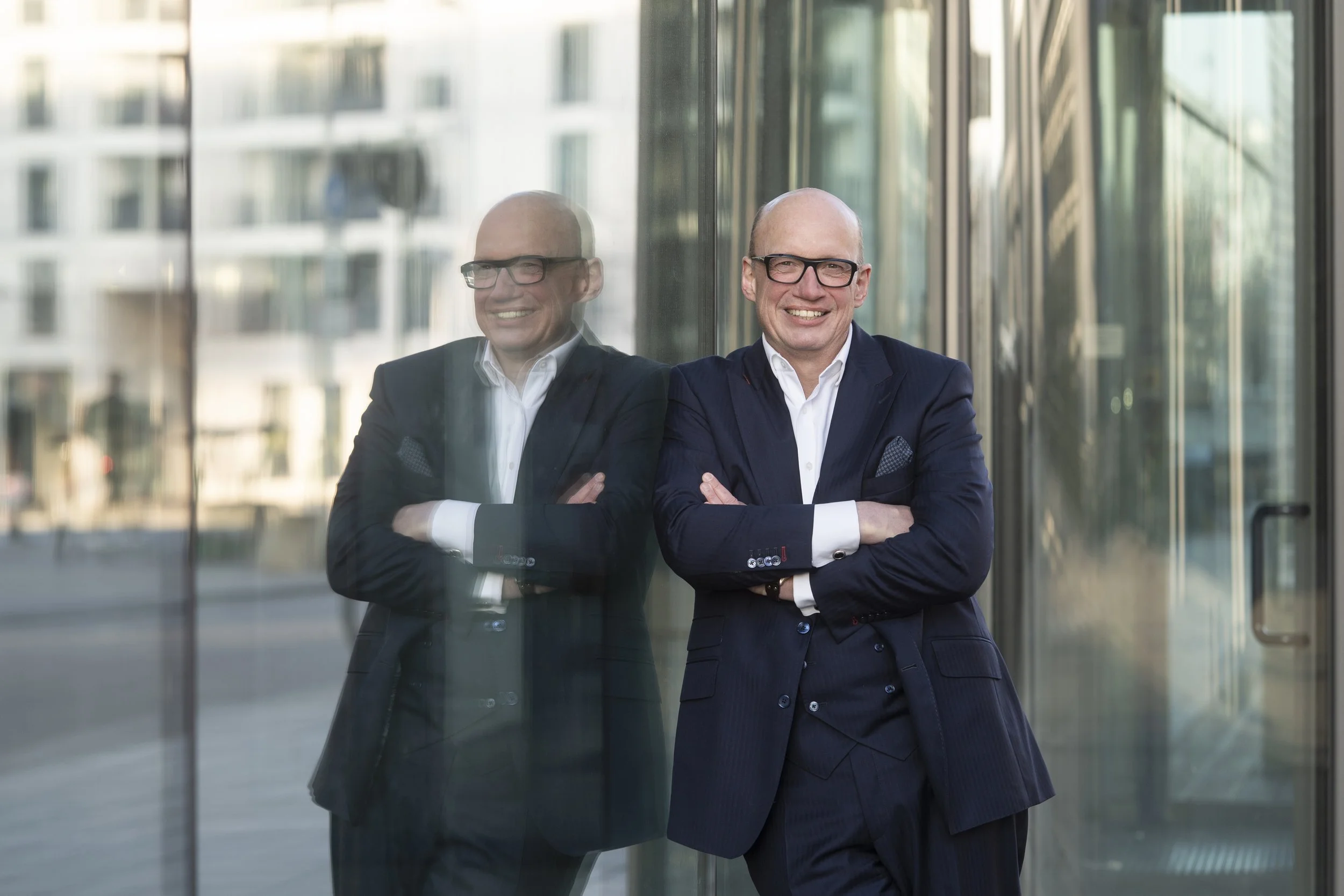 Ein Mann in Anzug steht vor einem modernen Glasgebäude, er lächelt und hat die Arme verschränkt. Seine Spiegelung ist im Fenster sichtbar.