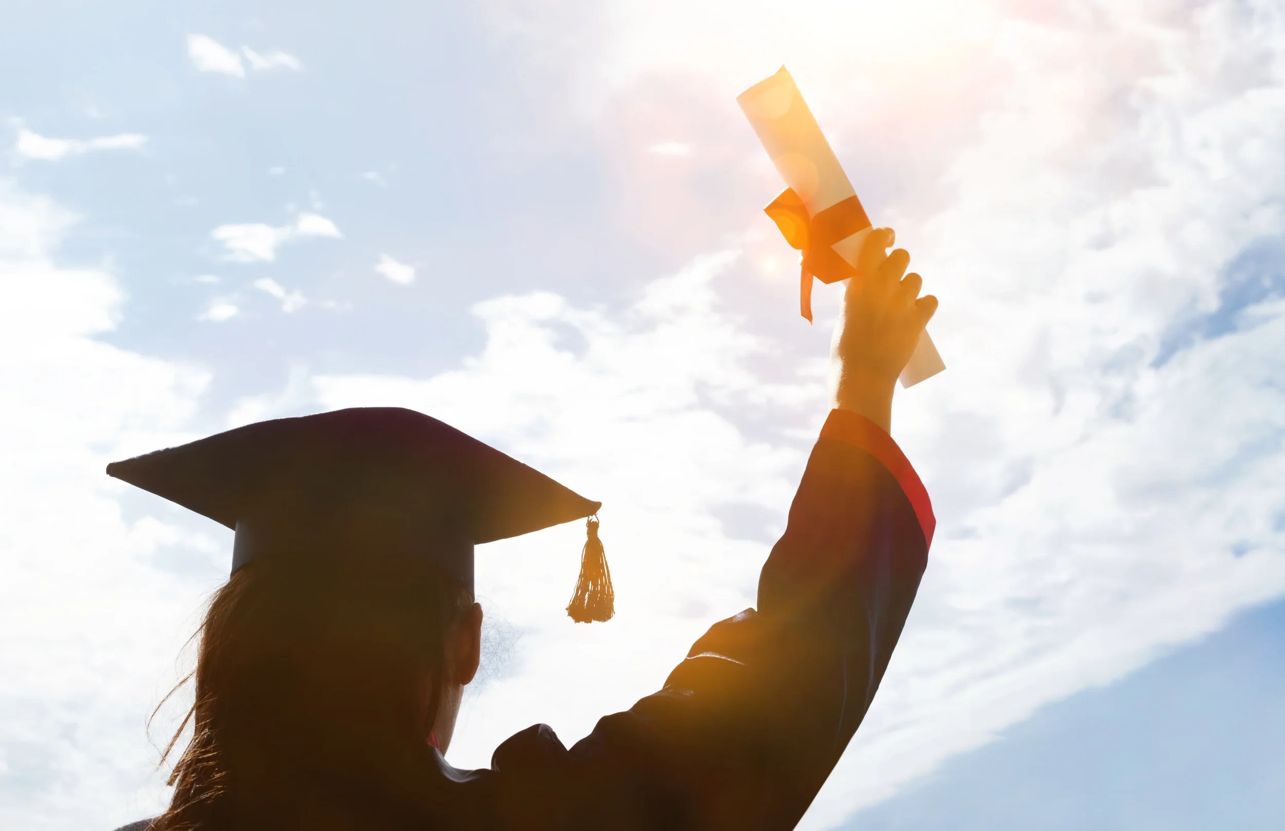 A person in a graduation cap holding a diploma paper against a bright, partly cloudy sky.