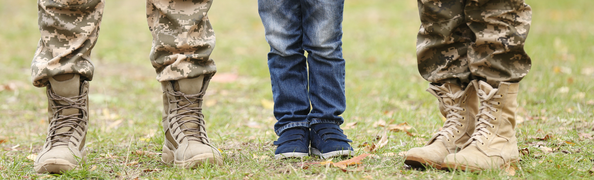 Close-up of three people's legs, two in military camouflage pants and boots, and one in regular jeans and sneakers, standing on grass outdoors.