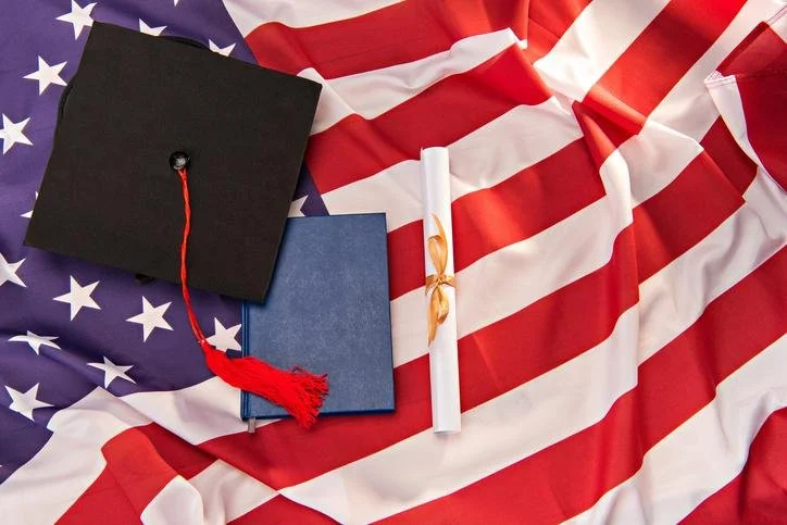A graduation cap, a diploma, and a blue book on an American flag background.