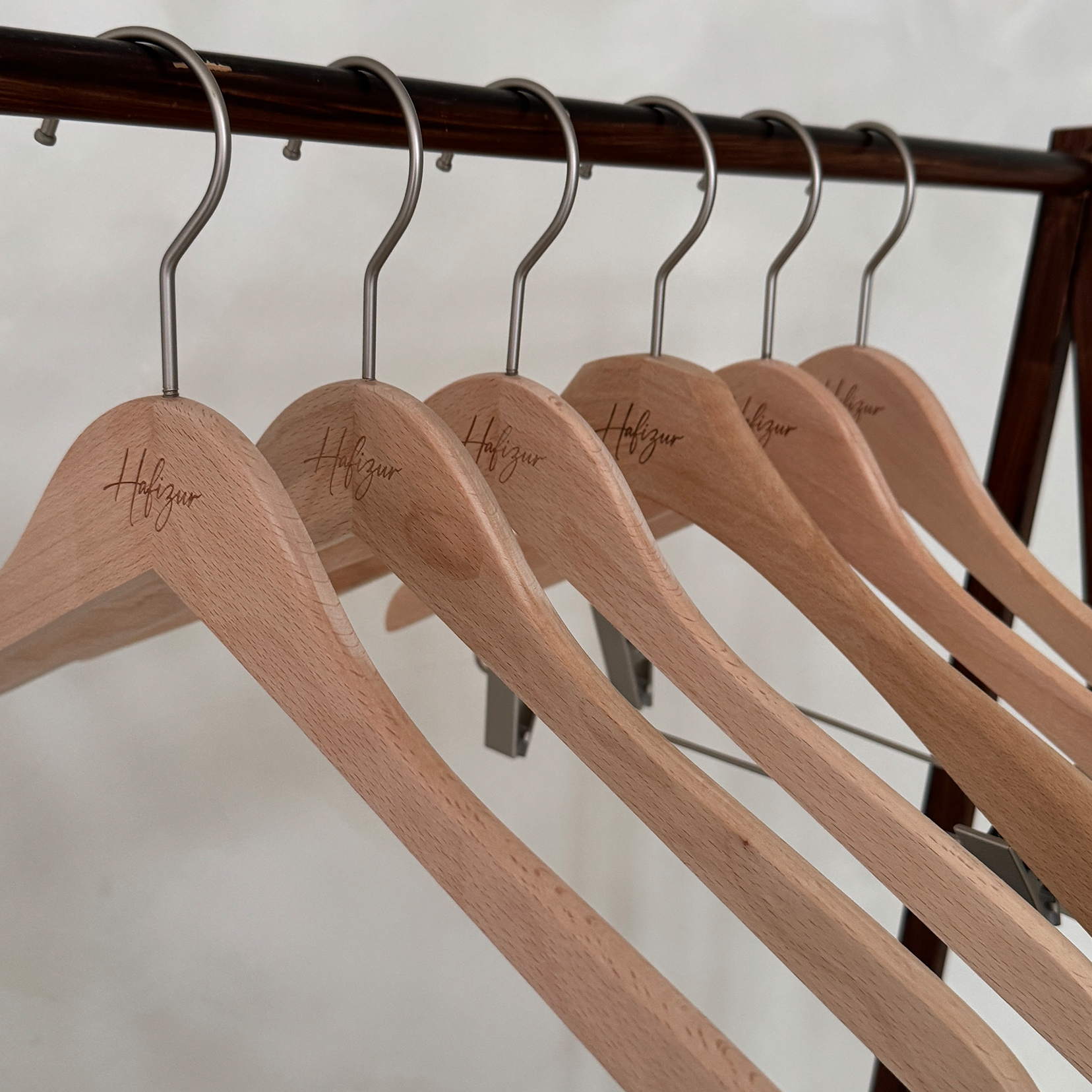 Six wooden hangers with metal hooks hanging on a wooden clothing rack, each hanger has the word Hafizur engraved on it.