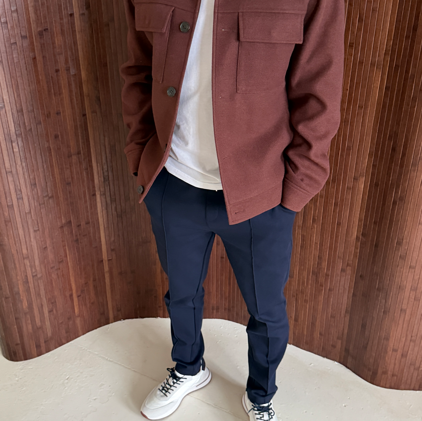 Person wearing a brown jacket, white t-shirt, dark blue pants, and white sneakers standing against a wooden background.