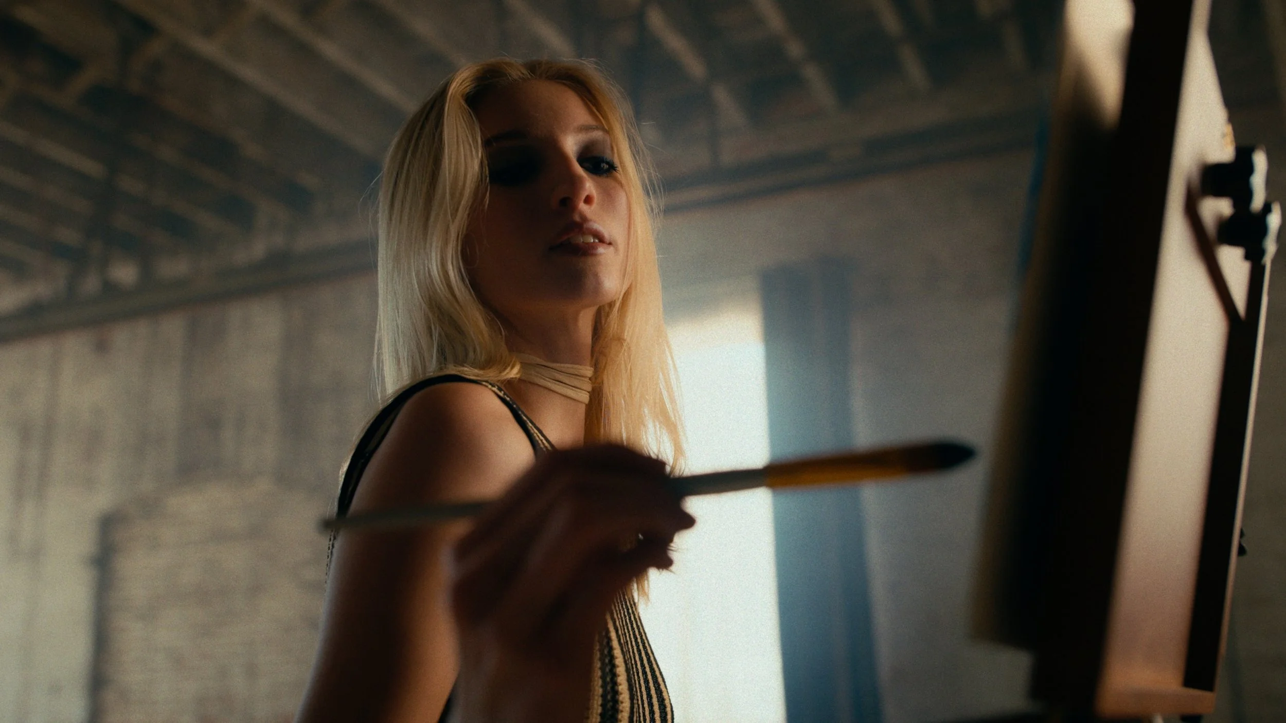 A woman with blonde hair painting on a canvas in a studio with an industrial ceiling and a soft backlight.