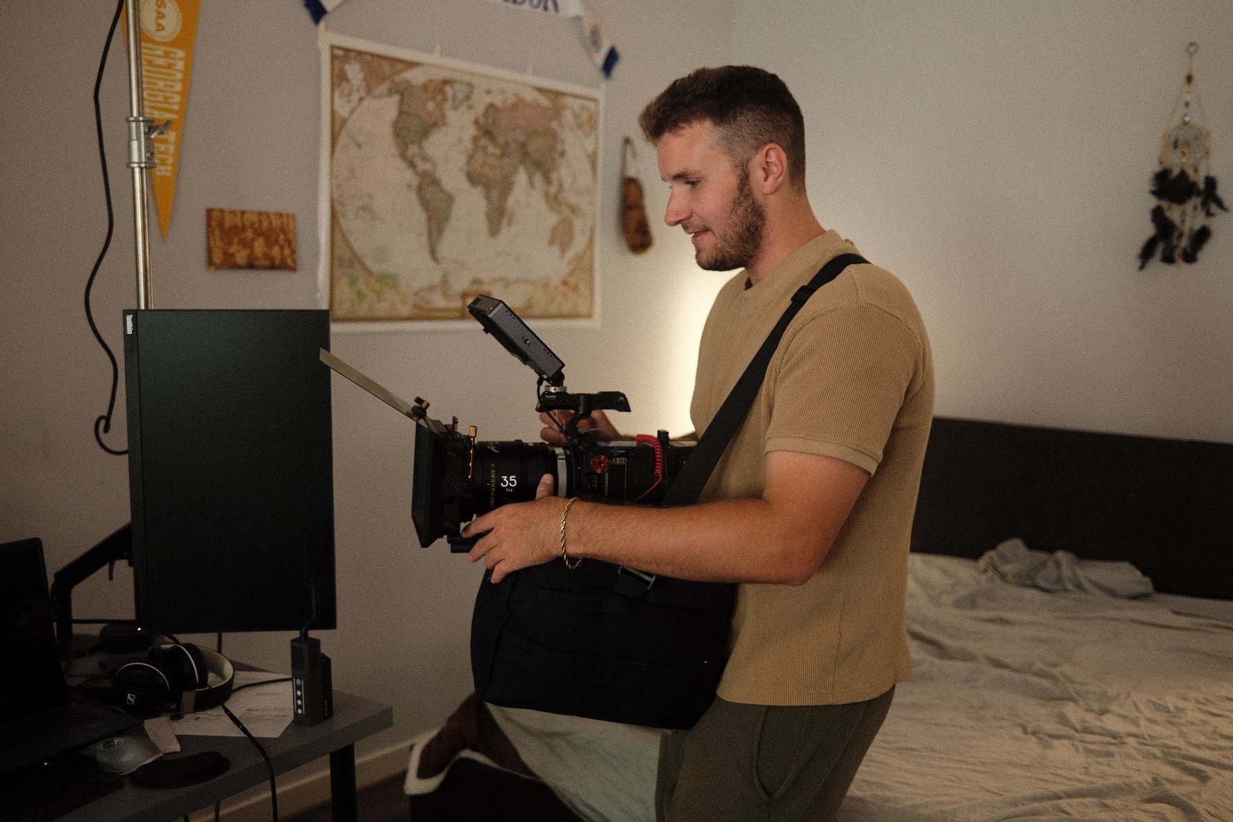 A man holding a professional video camera in a bedroom, with a large world map on the wall and a bed in the background.