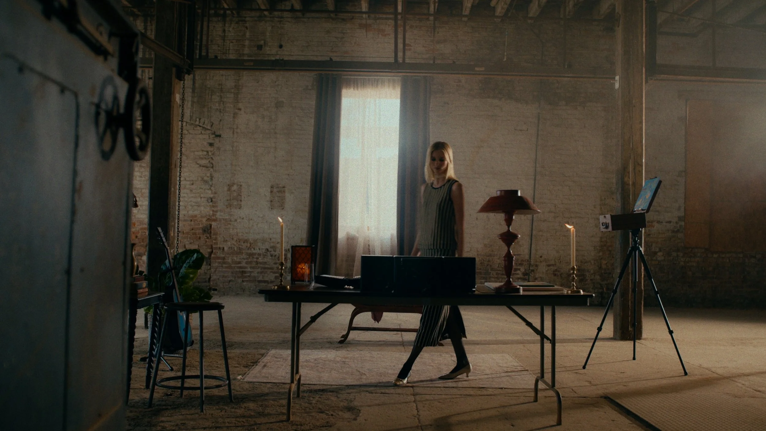 A woman with blonde hair standing behind a made-up table in a rustic, industrial-style room illuminated by a large window with sheer curtains. The table has a candle, a lamp, and a desktop computer, with a camera on a tripod nearby. The room has expo