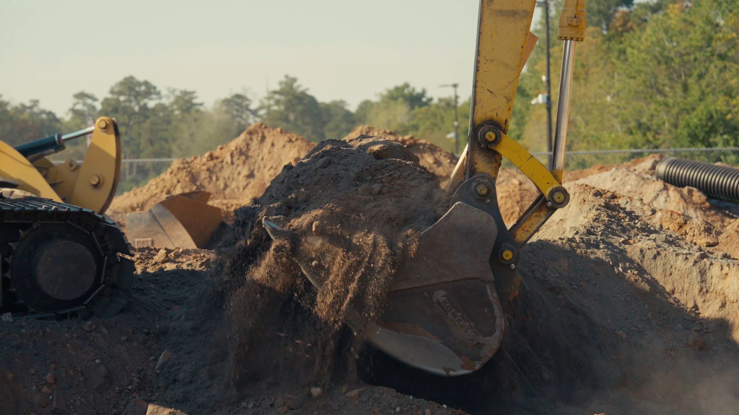 Close-up of a yellow excavator digging into dirt on an outdoor construction site, with piles of dirt and construction equipment in the background.