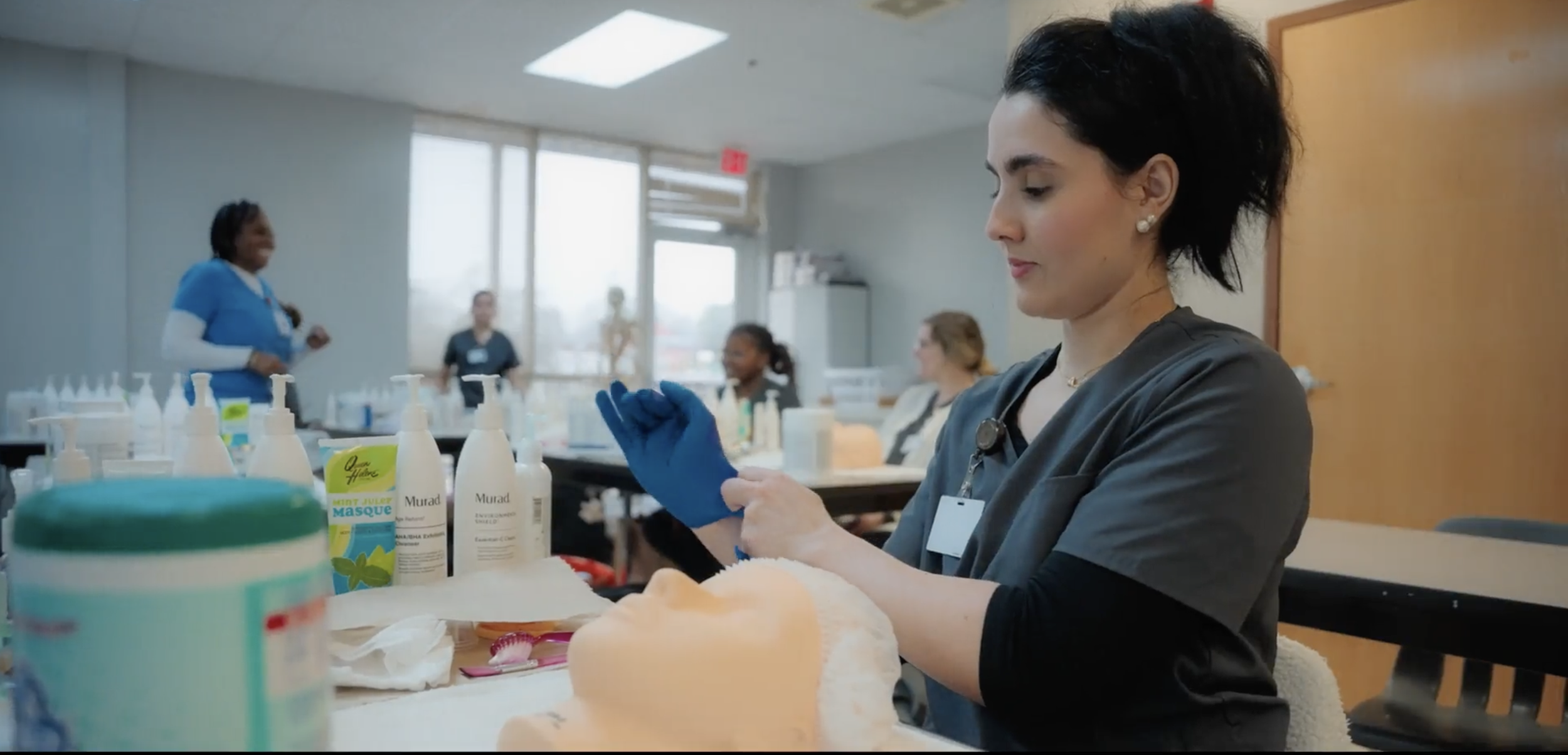Medical professional in scrubs and blue gloves preparing for a procedure in a classroom or training setting with other healthcare workers, medical supplies, and bottles on the table.