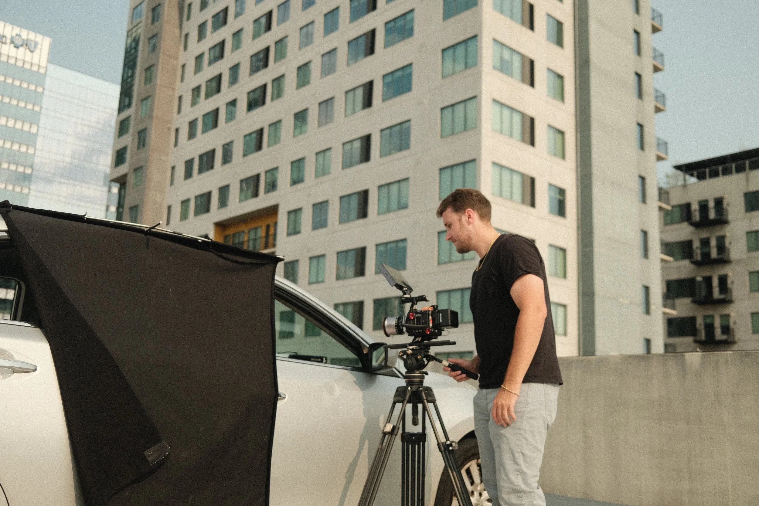 A man operating a camera on a tripod on an urban rooftop, with tall modern buildings in the background.