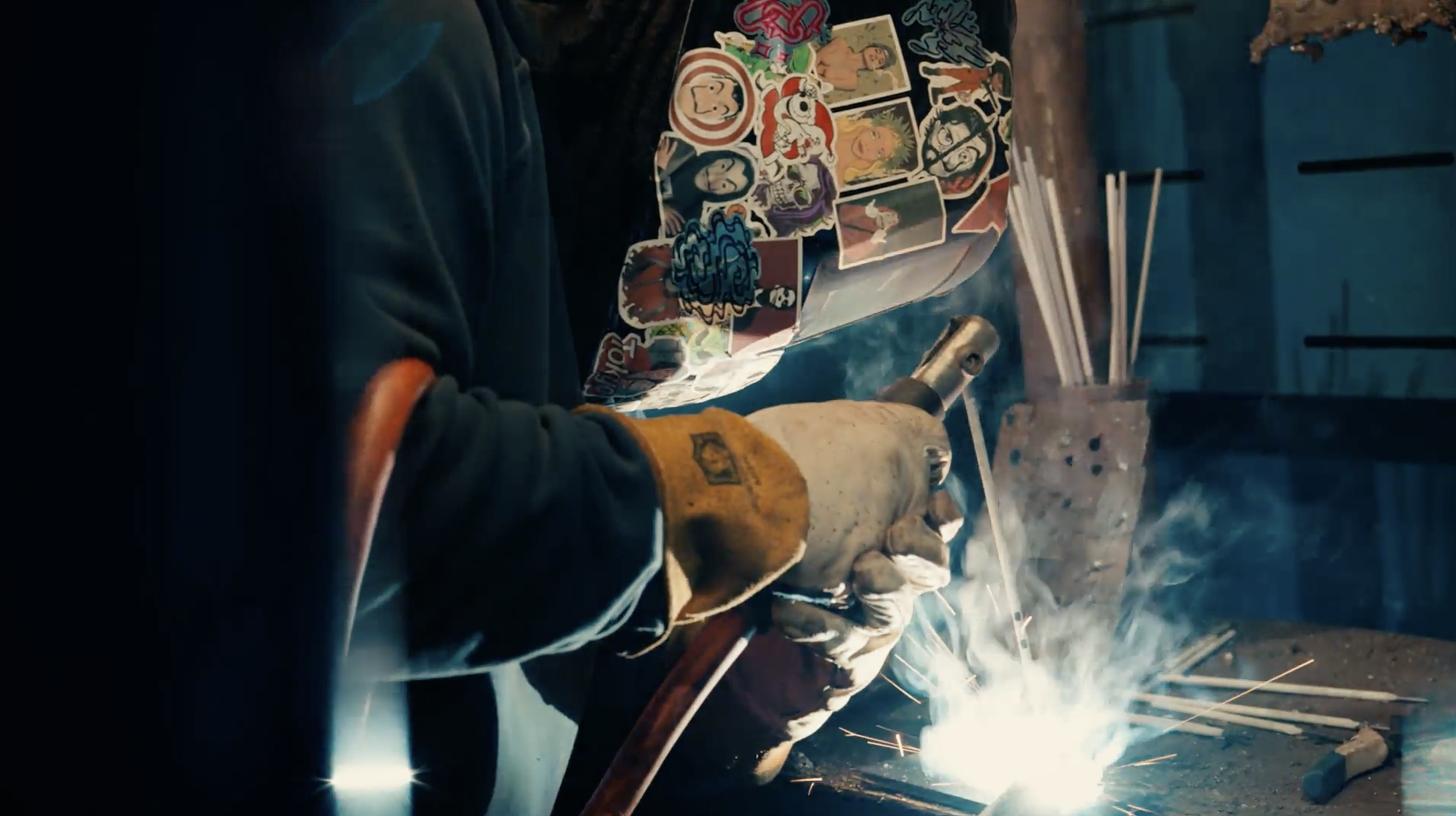 Person welding metal with sparks and smoke around, wearing protective gloves and clothing, workshop or industrial setting.