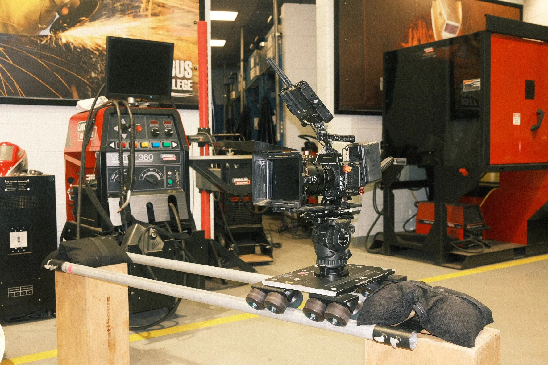 Film camera mounted on a dolly with wheels, positioned in front of a machine in a studio or workshop setting.