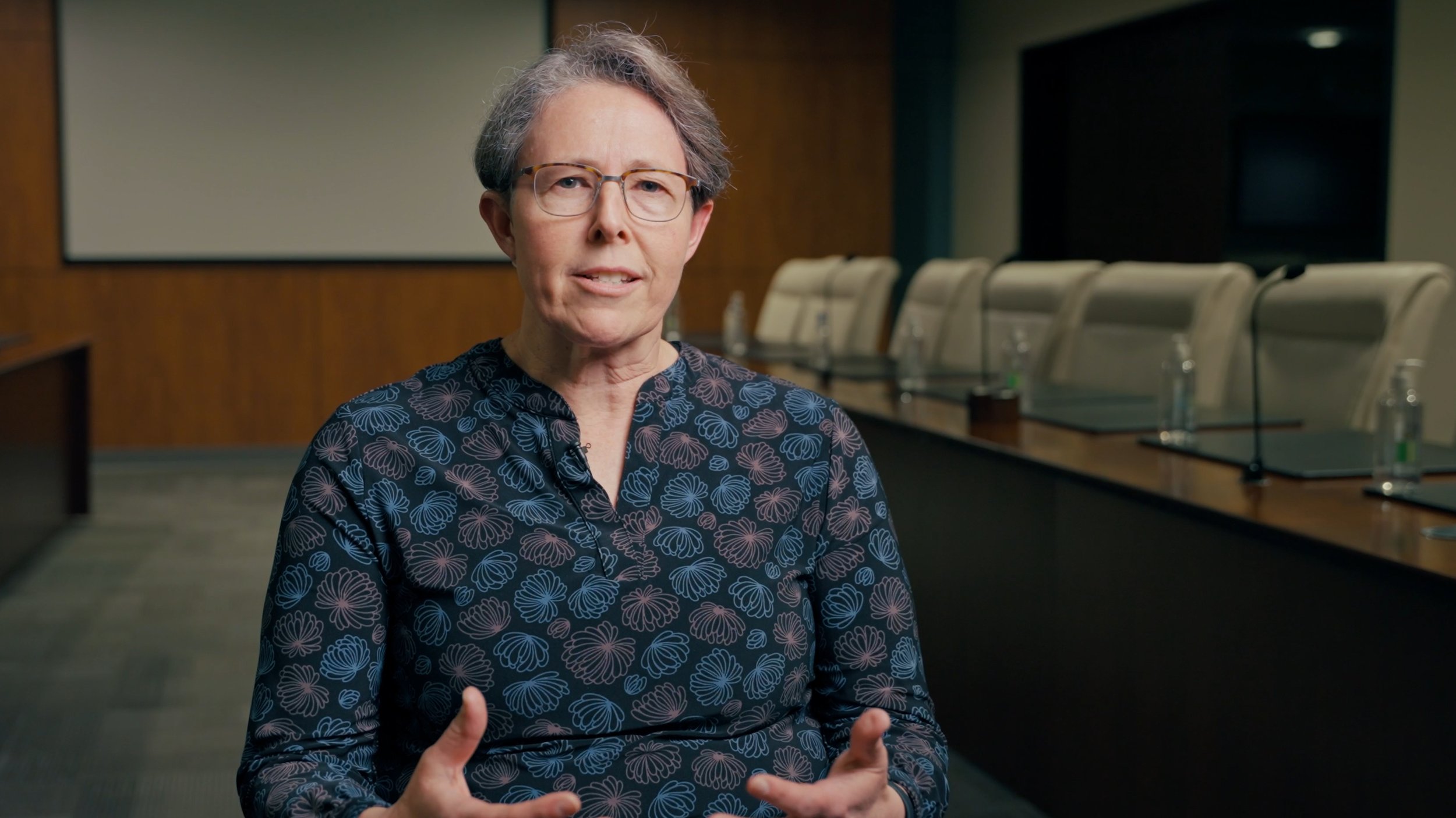 An older woman with short gray hair and glasses speaking in a conference room with a long table and beige chairs.