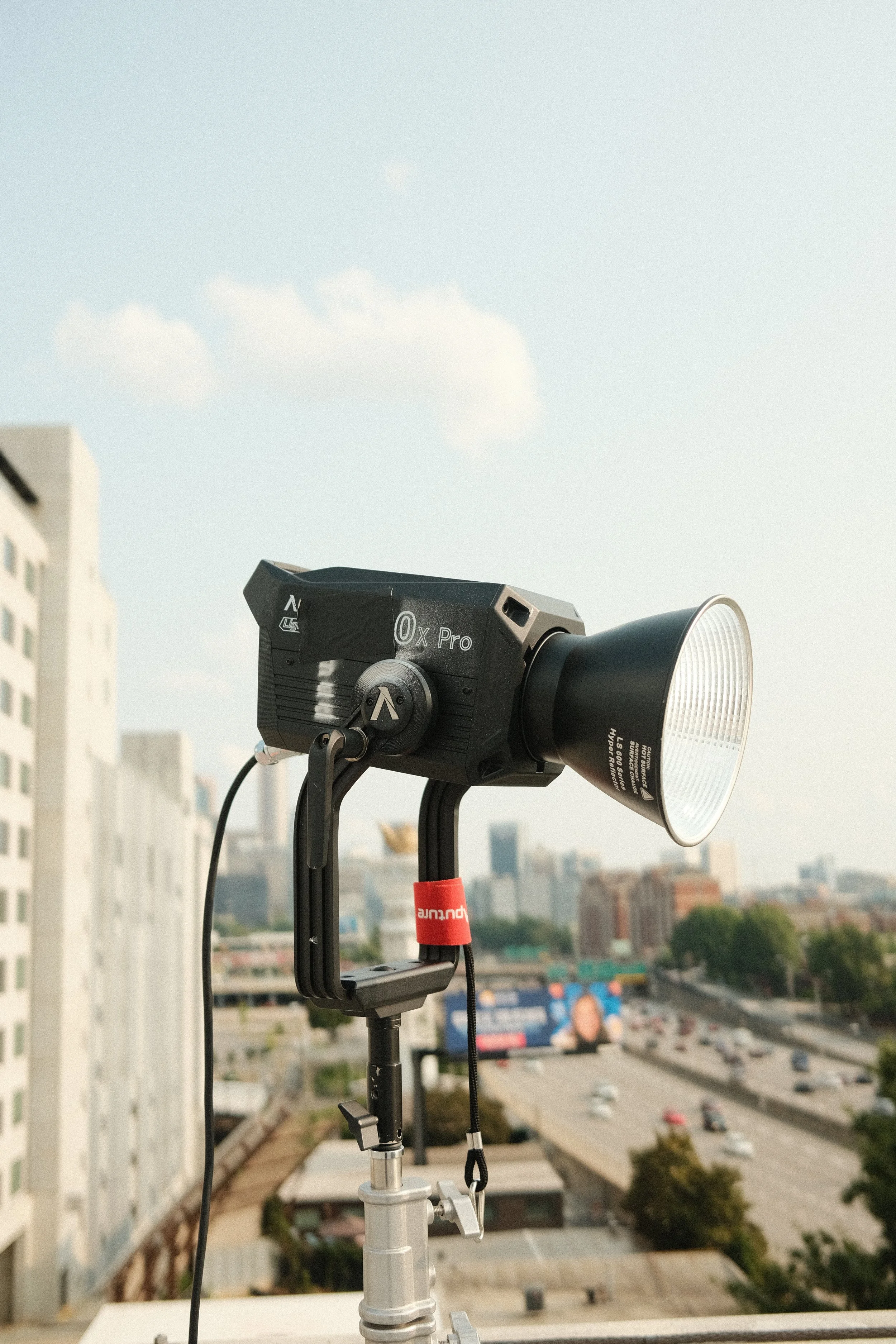 A professional studio light or spotlight mounted outdoors on a tripod, with a city skyline and highway in the background.