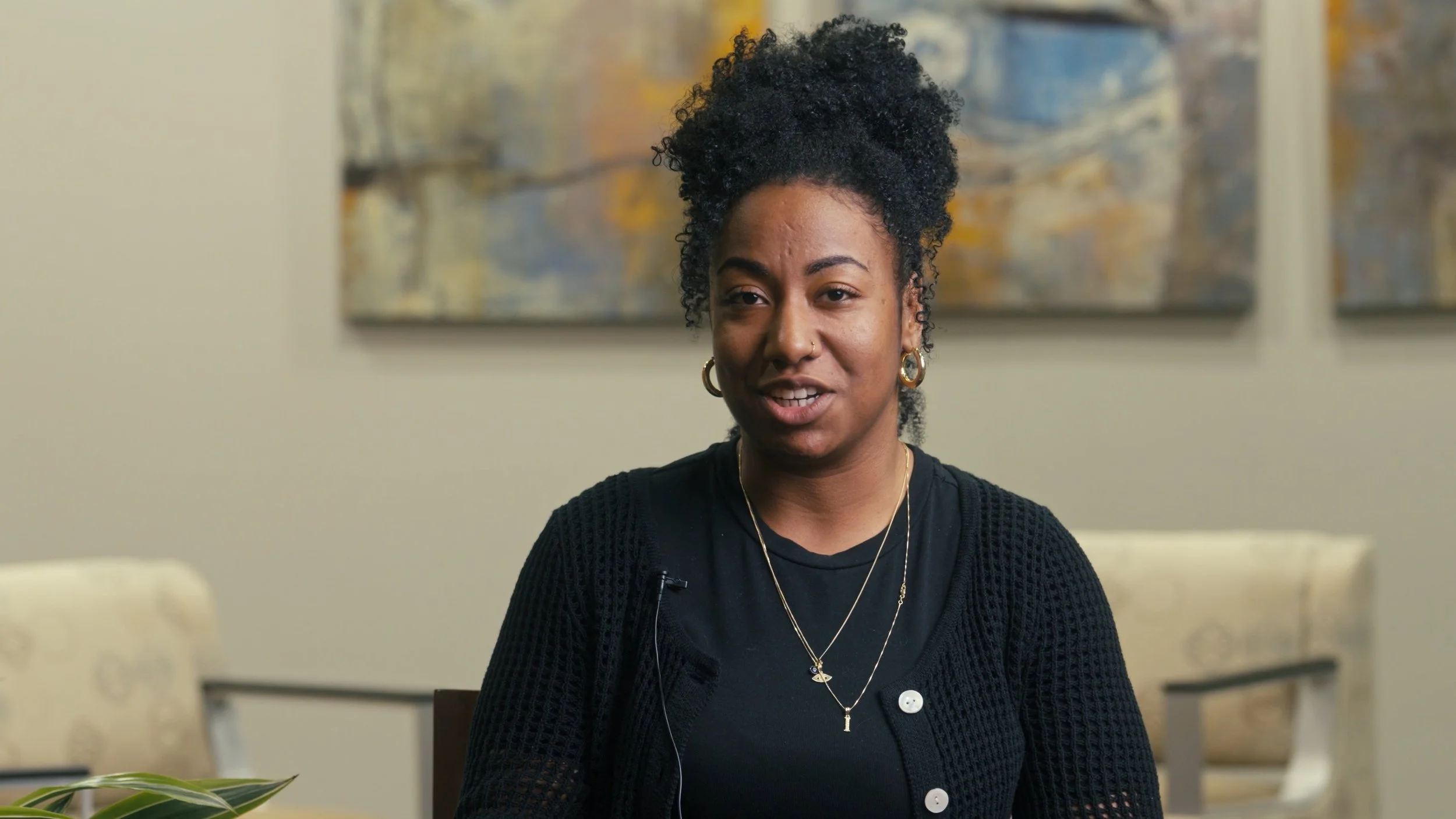 A woman with dark curly hair in a bun, wearing a black shirt and a black knitted cardigan, sitting at a table in a room with beige walls and abstract paintings in the background.