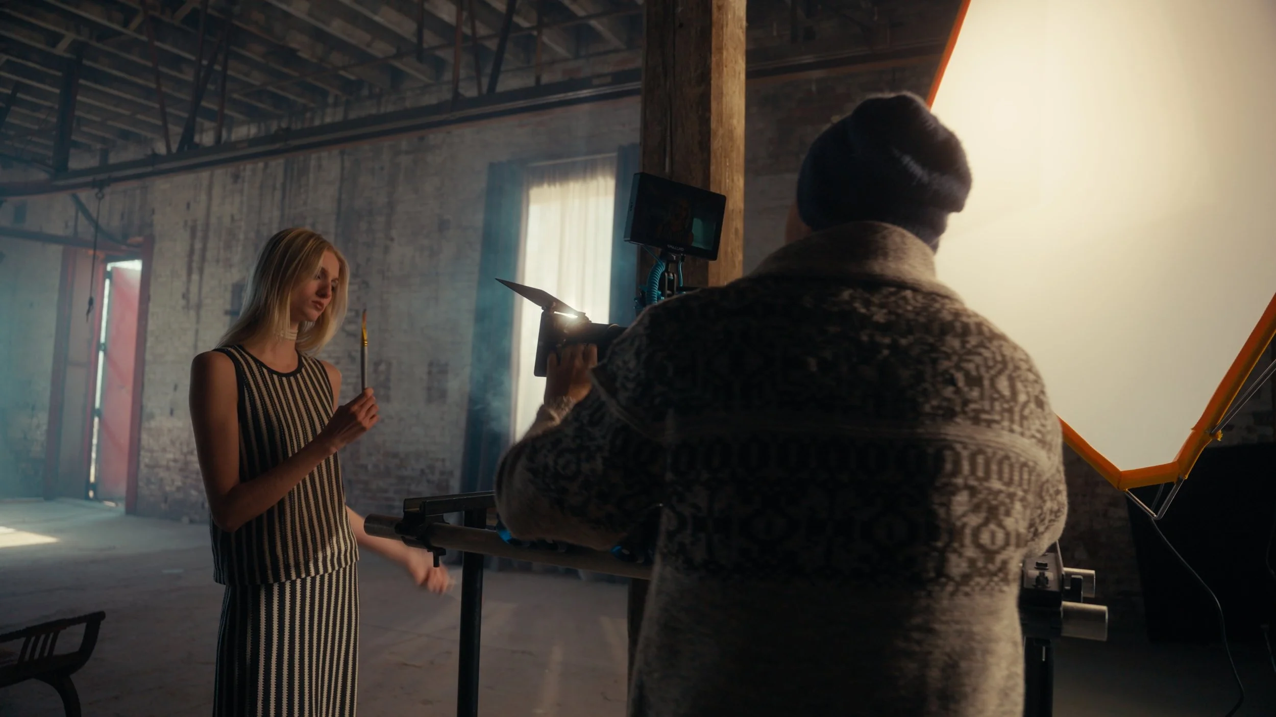 A female model with blonde hair in a black and white striped outfit during a photo shoot. The photographer is behind the camera, and a large reflector or lighting panel is on the right.