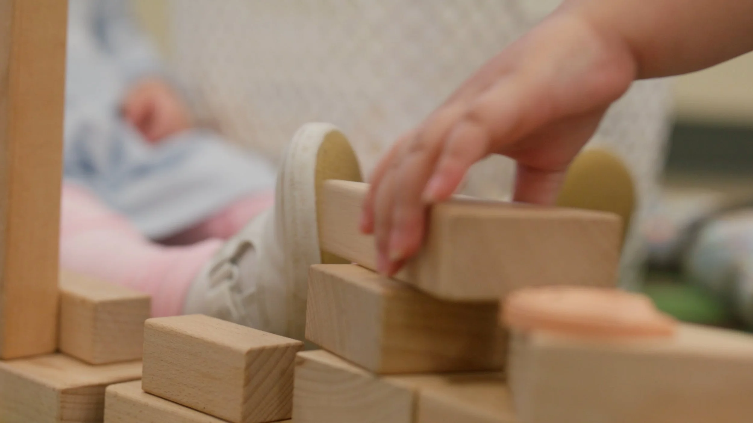 A child's hand placing a wooden block on a tower of wooden blocks, with a sleeping baby in the background.