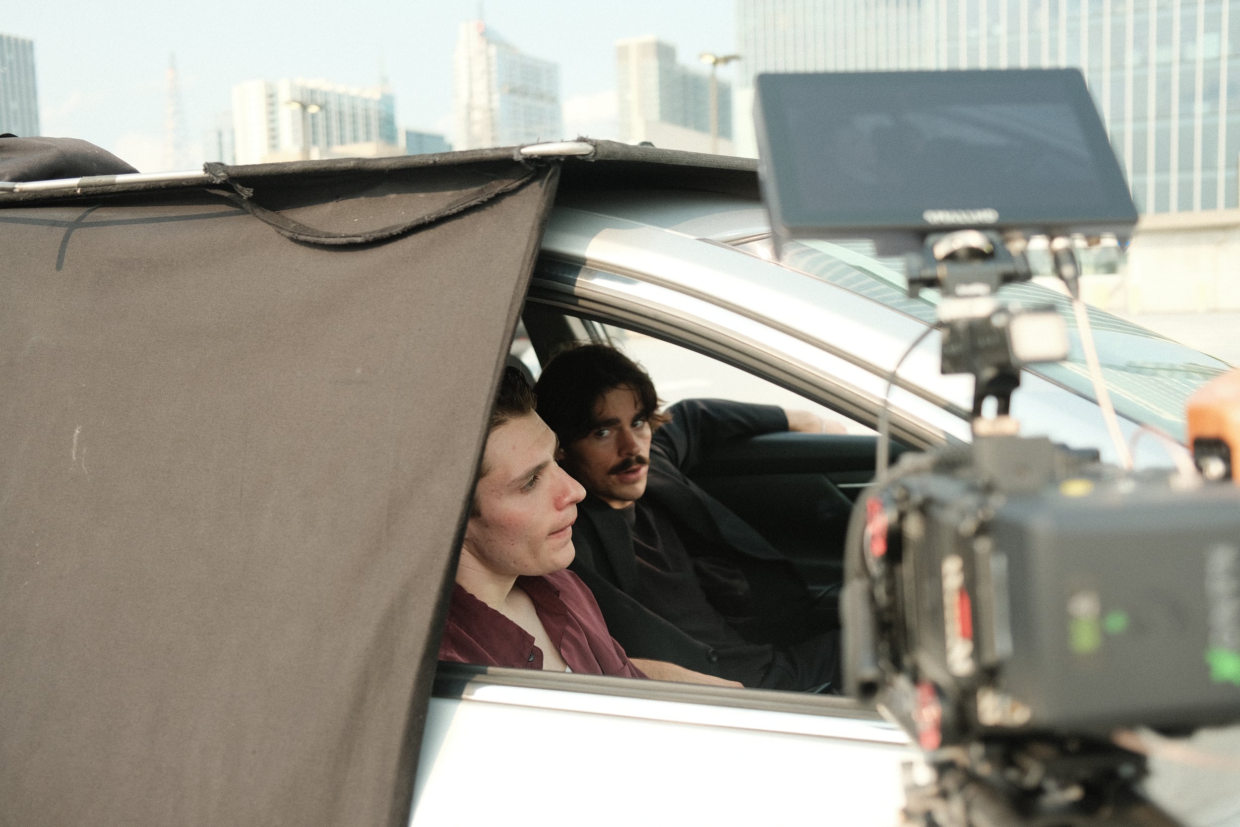 Two men sitting in a car behind a camera and filming equipment, with city buildings in the background.