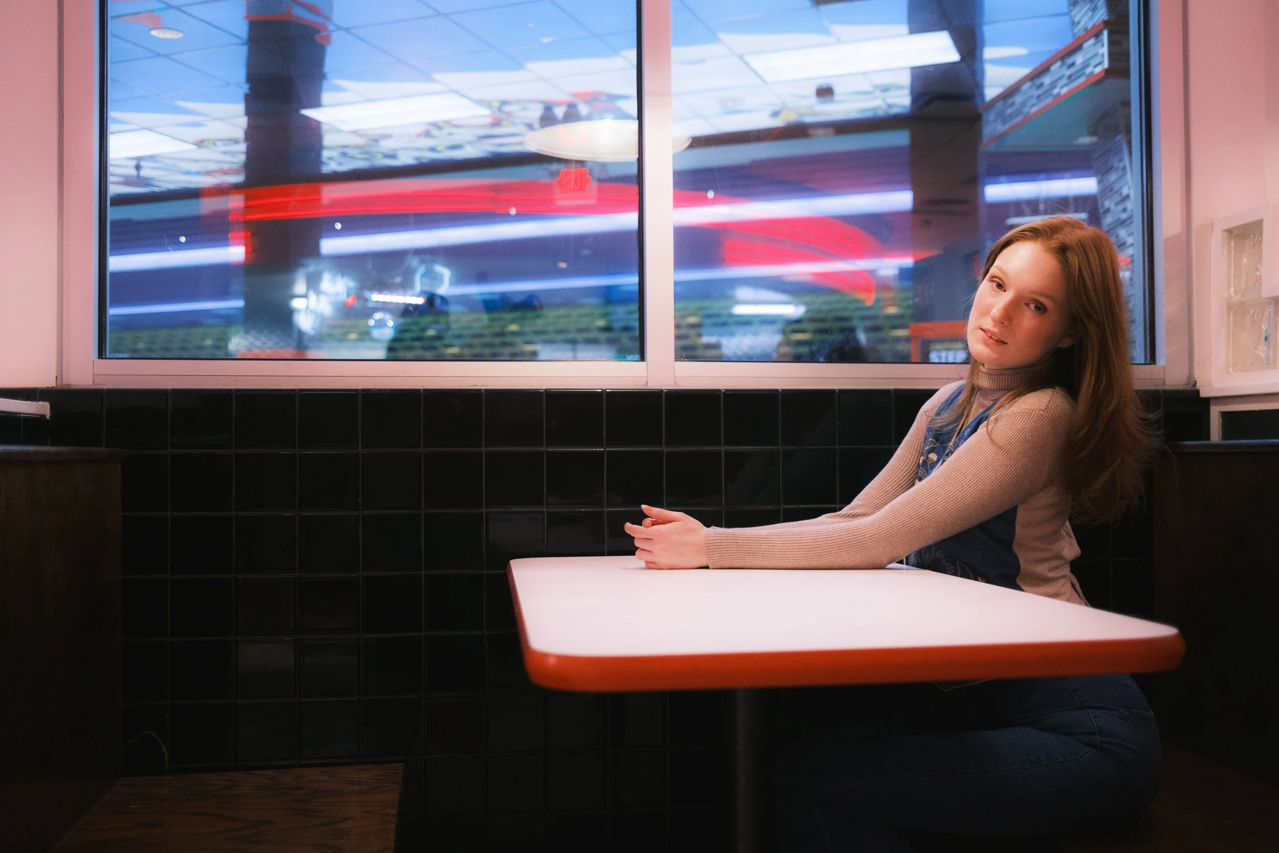 A young woman with red hair sitting alone at a booth in a restaurant or diner, looking towards the camera with a neutral expression, near a window with a cityscape visible outside at night.