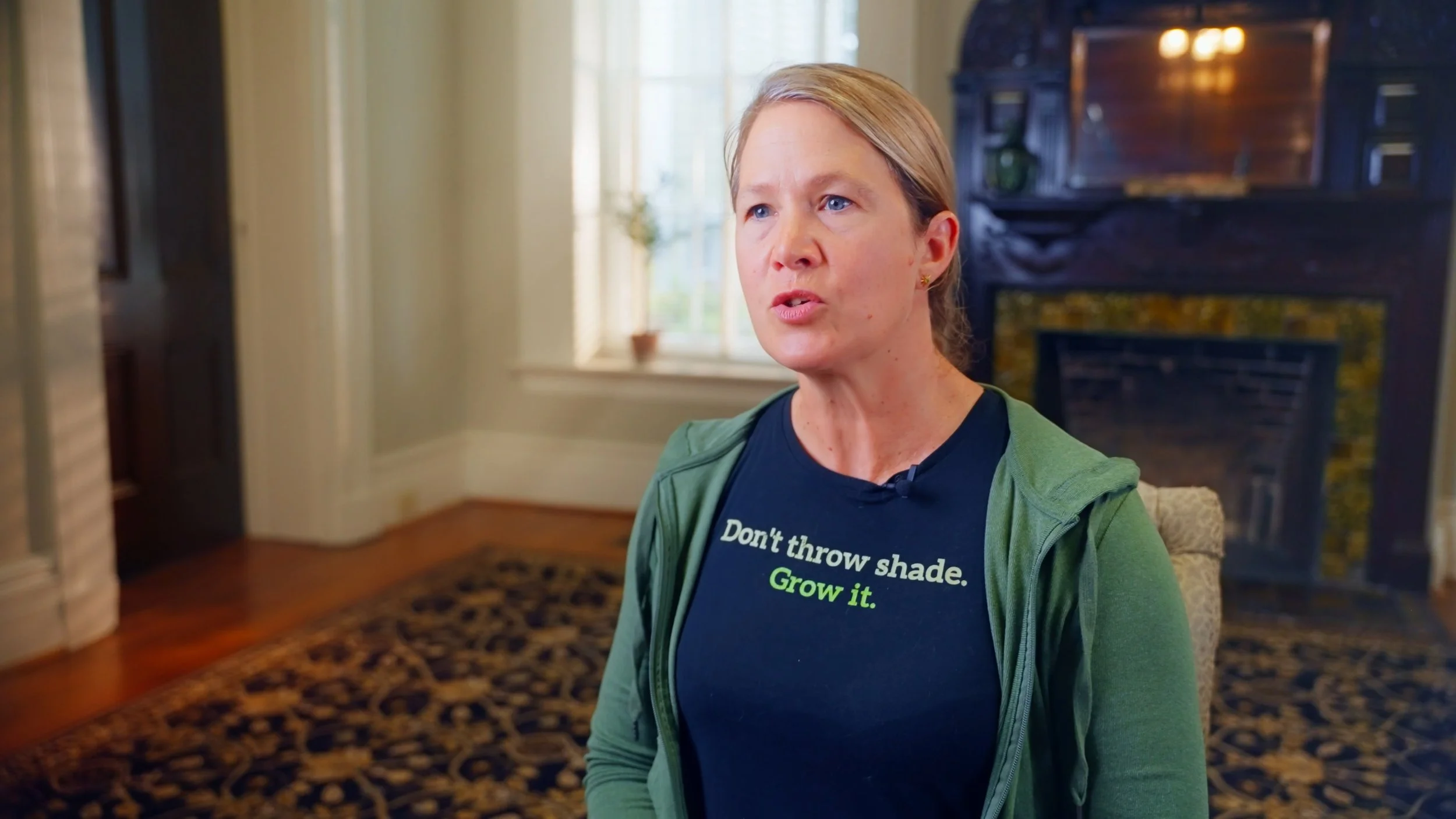 A woman with blonde hair and blue eyes sitting in a room with a fireplace and a window behind her, wearing a black T-shirt that says 'Don't throw shade. Grow it.' and a green zip-up jacket.