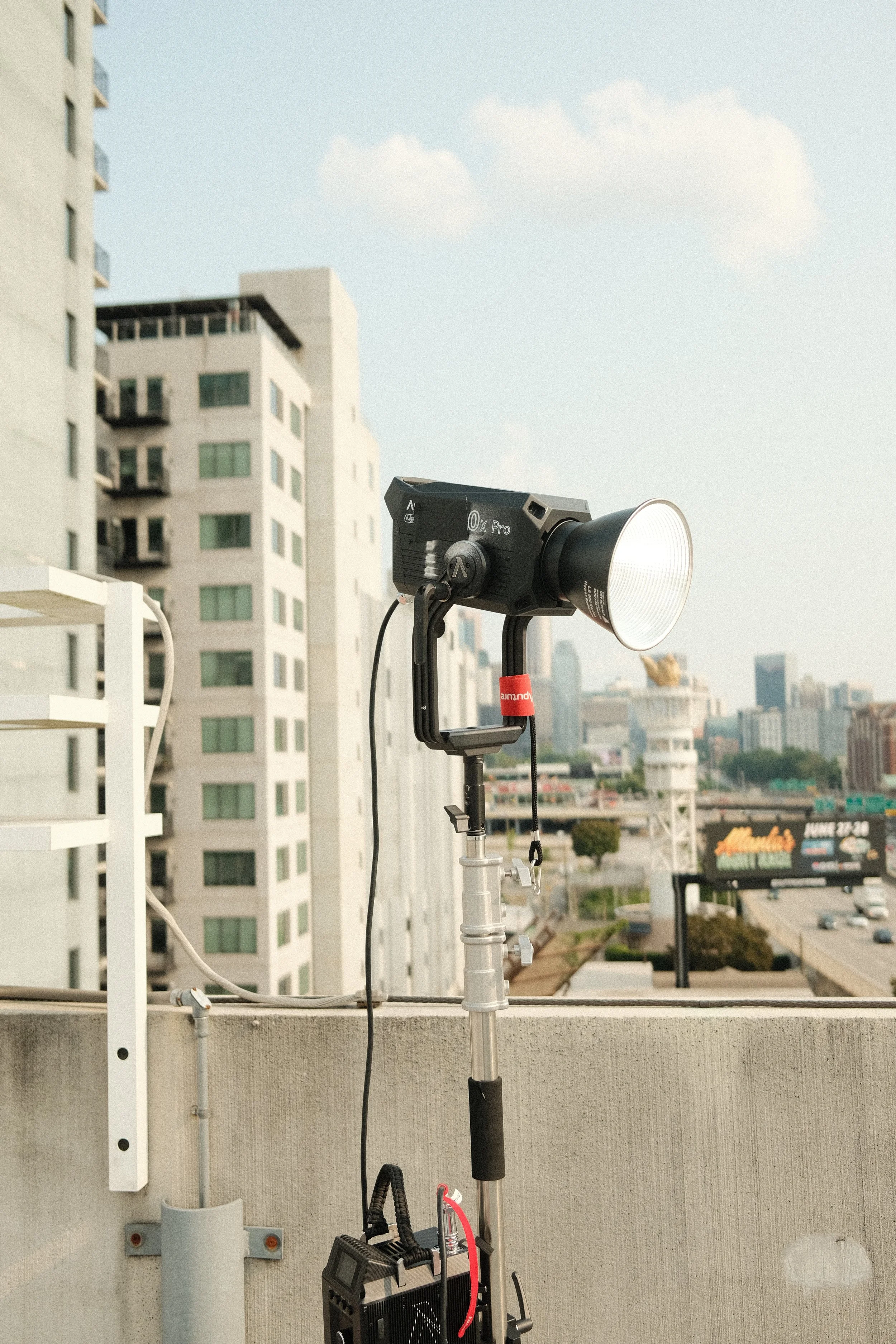 Photograph of a photography lighting setup on a city rooftop, featuring a large black studio light mounted on a tripod, with a cityscape of tall buildings and a highway in the background.