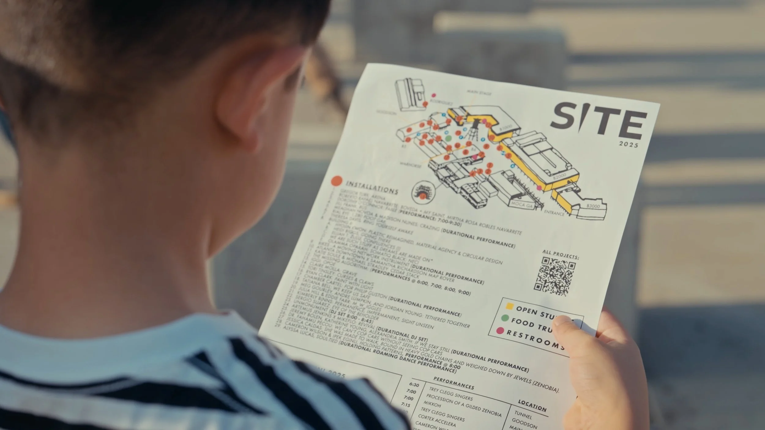 Person holding a site map with a layout of installations, open studios, food trucks, and restrooms, including color-coded legend and QR code.