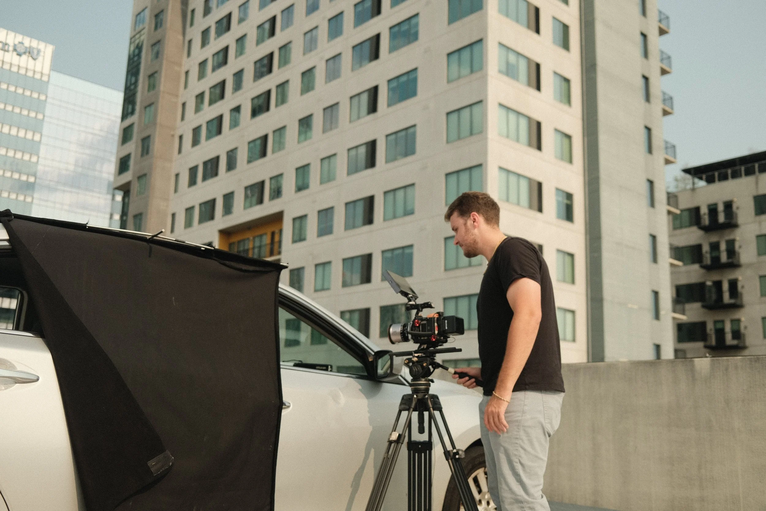 A man operating a camera on a tripod after filming a white car with a black screen in an urban area with tall modern buildings.