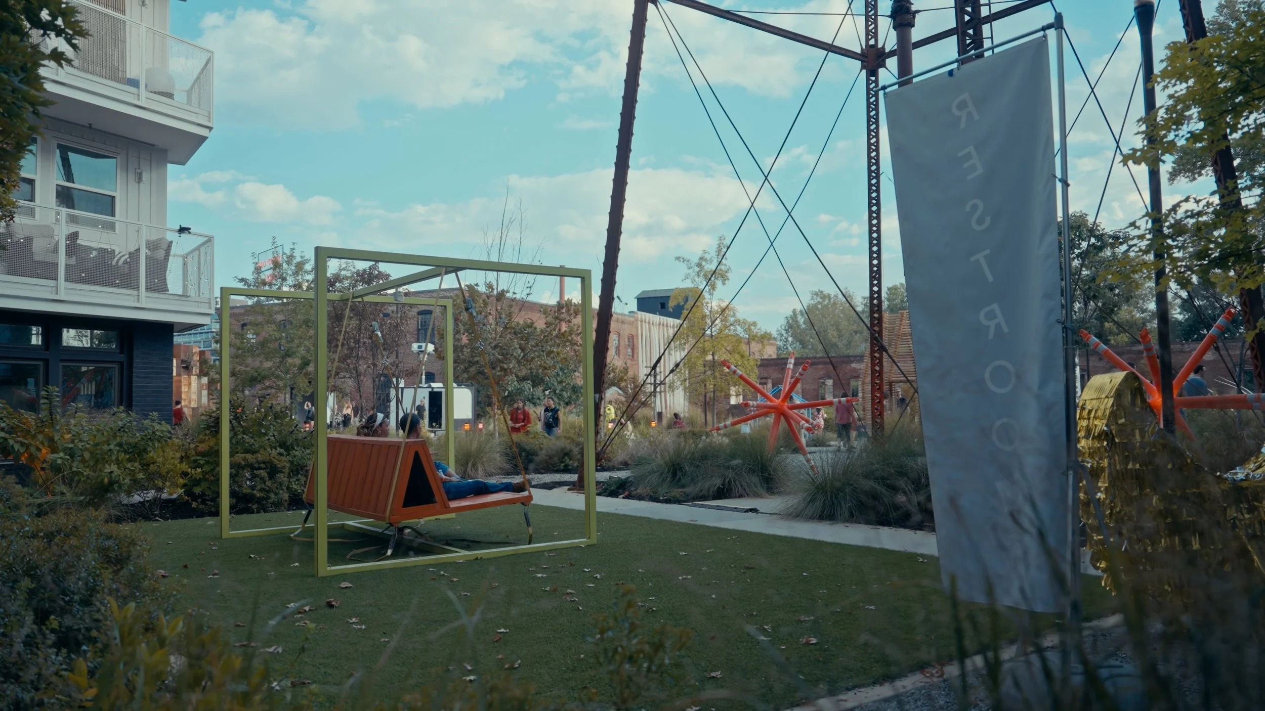 Urban park with modern outdoor furniture, some people walking in the background, and artistic sculptures, with a tall banner displaying text, under a partly cloudy sky.