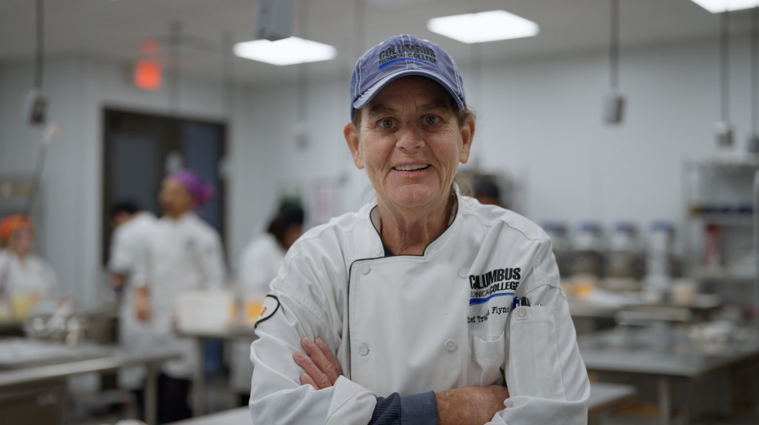Senior female chef wearing a white chef coat with Columbus Technical College logo standing with arms folded in a commercial kitchen.